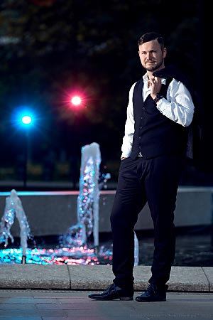 A young man wearing a suit is standing in front of a fountain. Behind him there are two coloured lights.