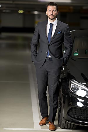 A young man wearing a suit is standing beside his car.